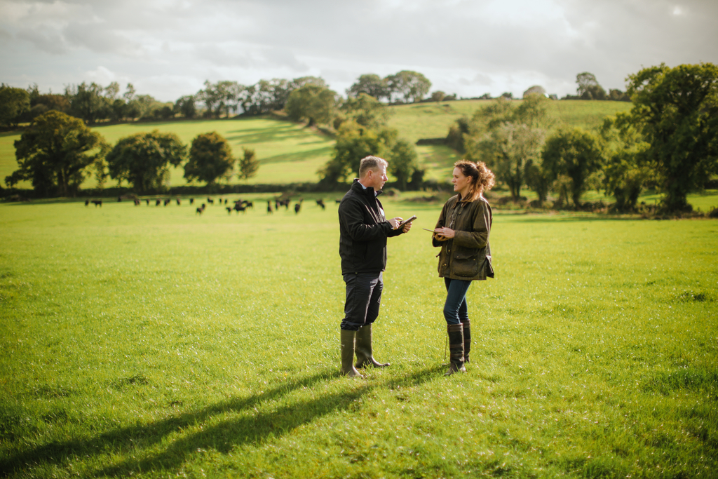Man and woman standing in green field talking to each other, trees and sky in background